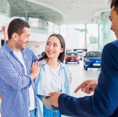 A car dealership scene featuring a salesperson engaging with two customers while showcasing vehicles in the background.